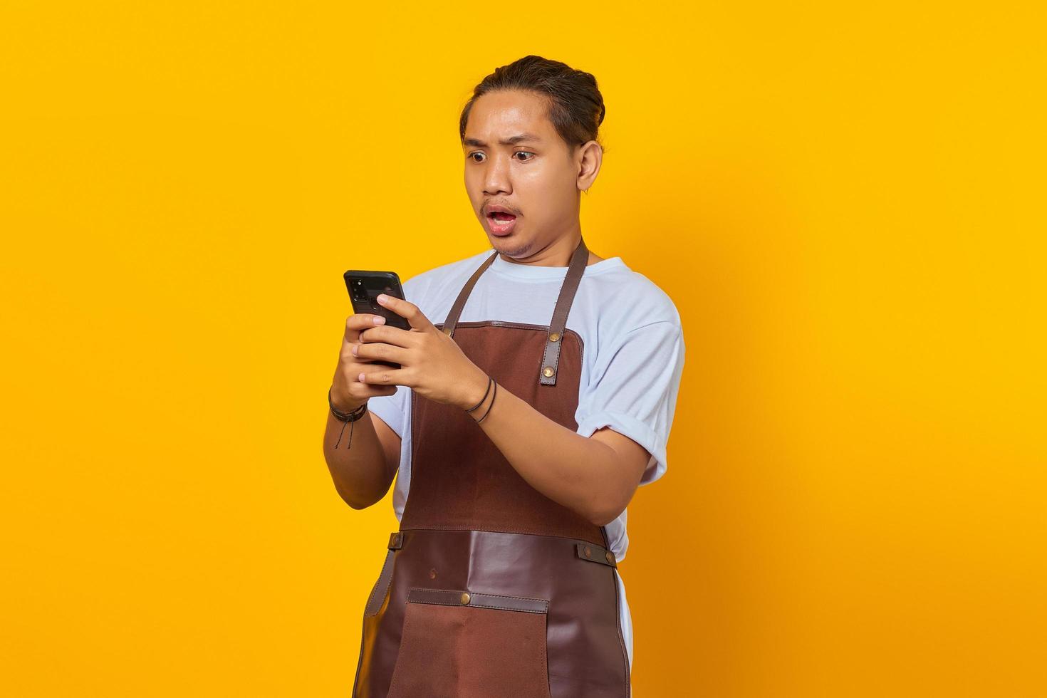 Retrato de guapo barista que está conmocionado y sorprendido al ver la promoción de descuento en el teléfono inteligente sobre fondo amarillo foto