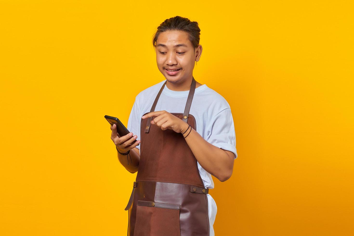 Portrait of Asian young man wearing apron surprised to see incoming message on smartphone on yellow background photo