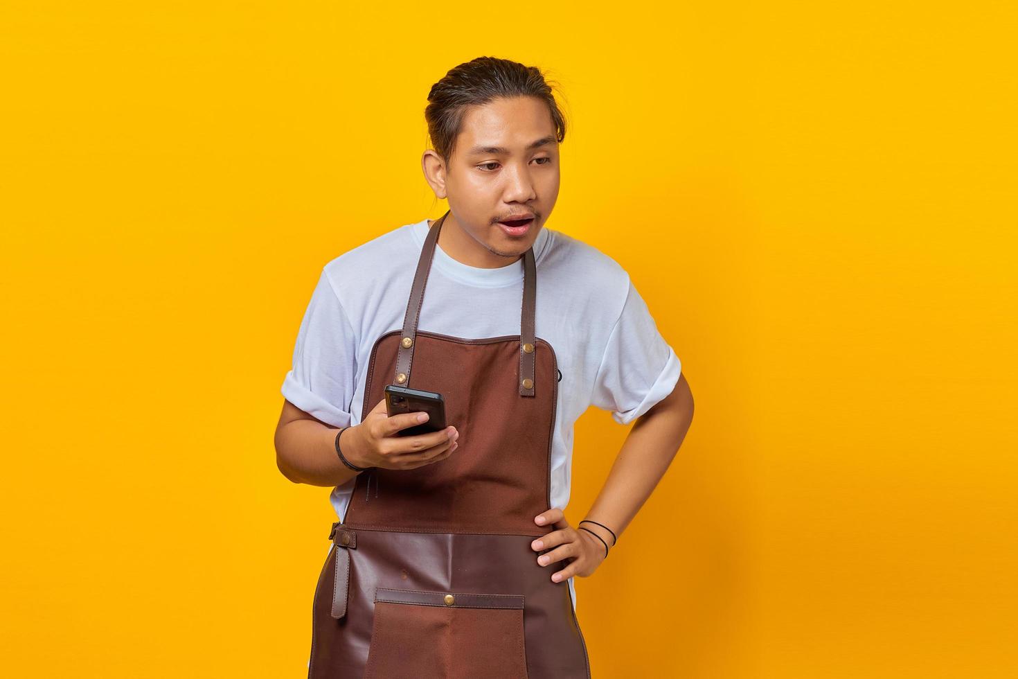 Portrait of Asian young man wearing apron surprised to see incoming message on smartphone on yellow background photo