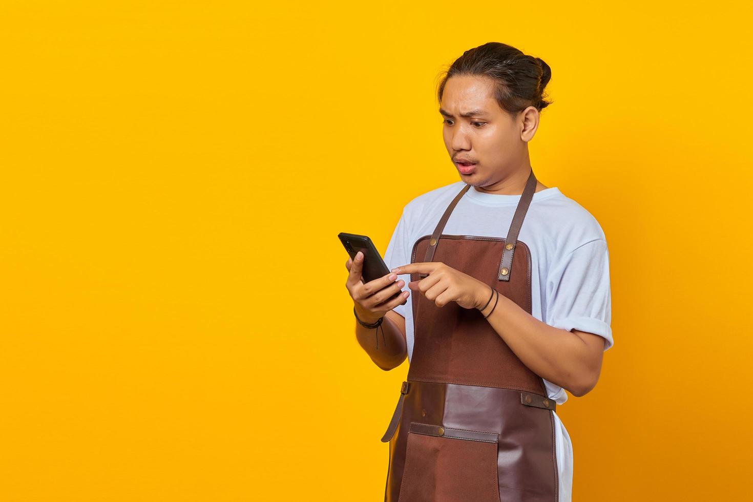 Portrait of Asian young man wearing apron surprised to see incoming message on smartphone on yellow background photo
