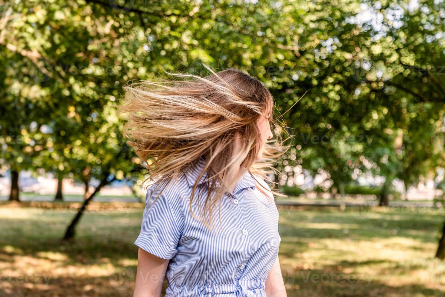 Joven mujer rubia caucásica en un vestido de verano azul despojado sacudiendo su cabello en el parque al aire libre en un día soleado foto