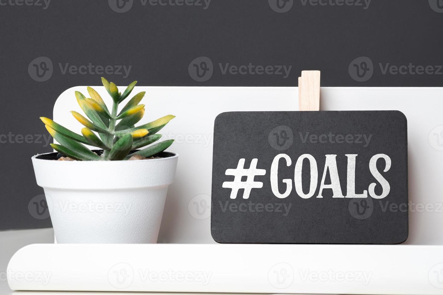 Goals on blackboard on pencil box and green plant in white pot on table and dark grey wall in office desk photo