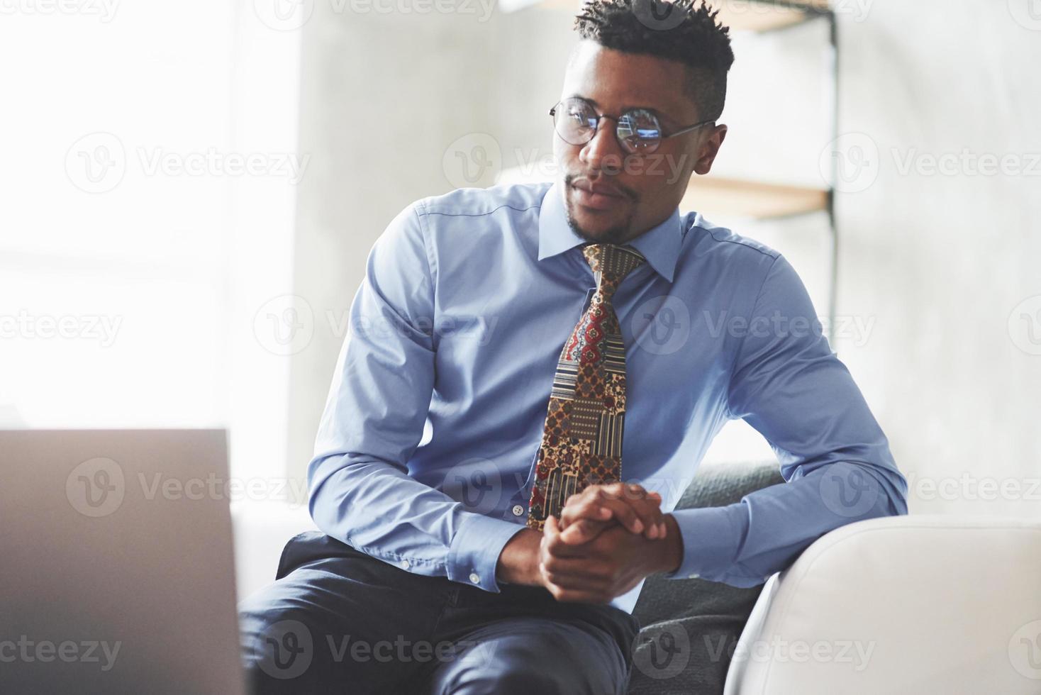 Photo of young stylish black man in the suit and glasses sitting on the sofa and looking at a laptop