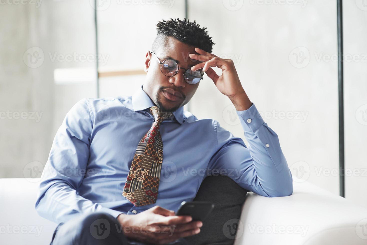 descansando en la oficina. Foto de joven negro elegante en traje y gafas sosteniendo y mirando el teléfono mientras está sentado en el sofá