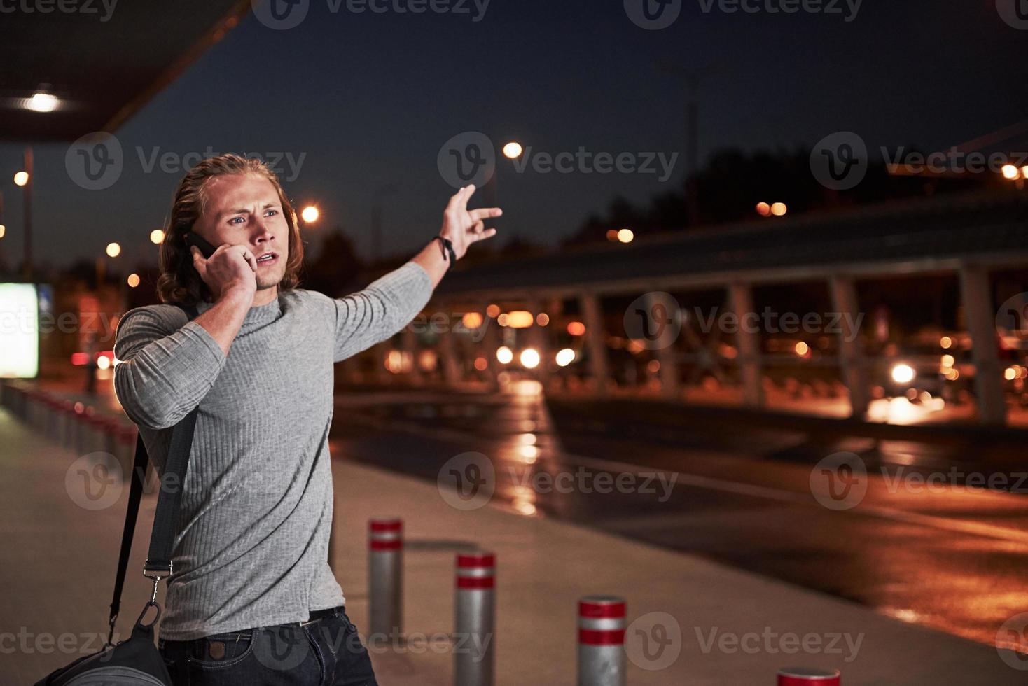 Active hand gestures. Young stylish man walking through the night street near the road and having a conversation on the phone photo