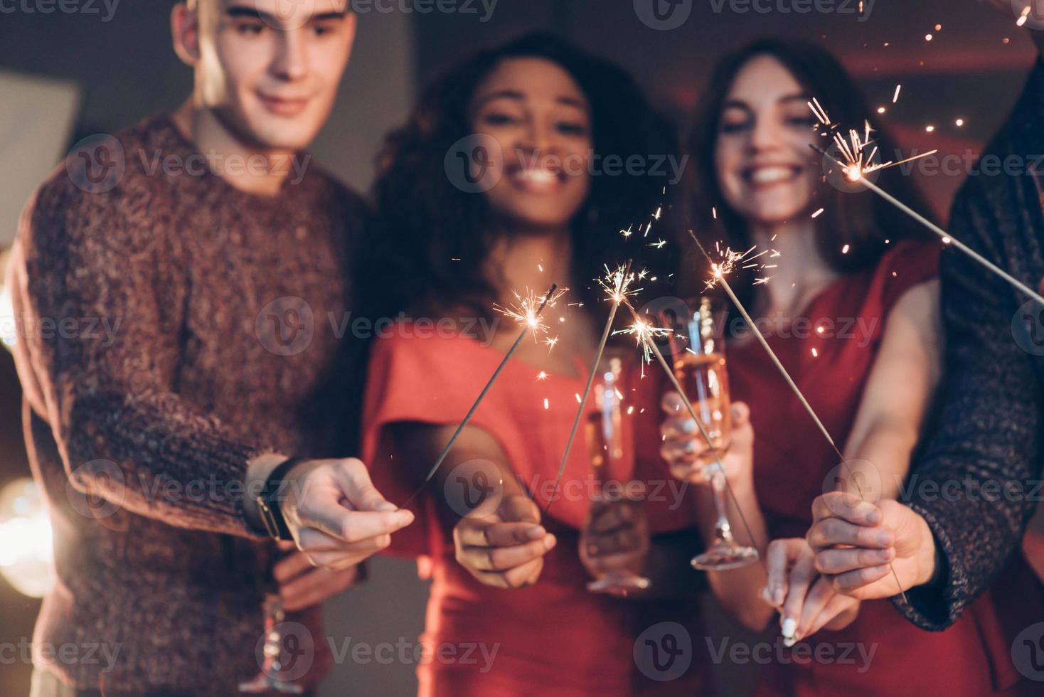sonreír y sentirse feliz. amigos multirraciales celebran el año nuevo y sostienen luces de bengala y vasos con bebida foto