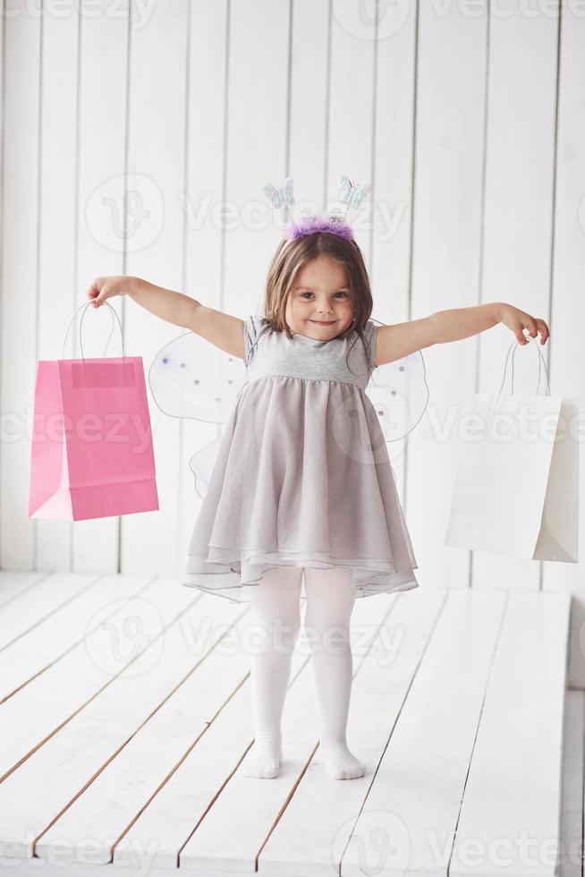 sosteniendo paquetes de papel mientras está de pie en el piso de madera blanca. hermosa niña con disfraz de hada divirtiéndose posando para las fotos