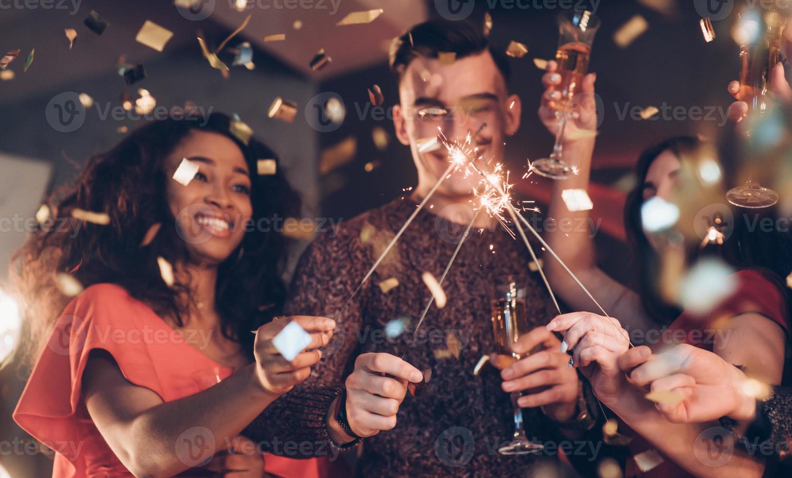 emociones sinceras. amigos multirraciales celebran el año nuevo y sostienen luces de bengala y vasos con bebida foto