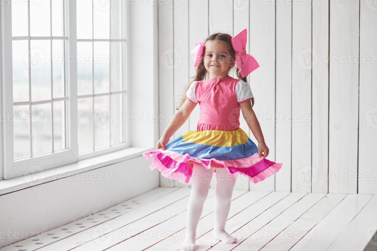 esta chica tiene un lindo vestido. Niño de pie junto a las ventanas en la habitación blanca y posando para la foto