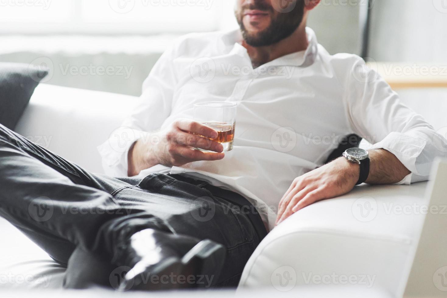 tener un merecido descanso. Foto recortada del joven empresario en ropa clásica sentado en el sofá con vaso de whisky en la mano