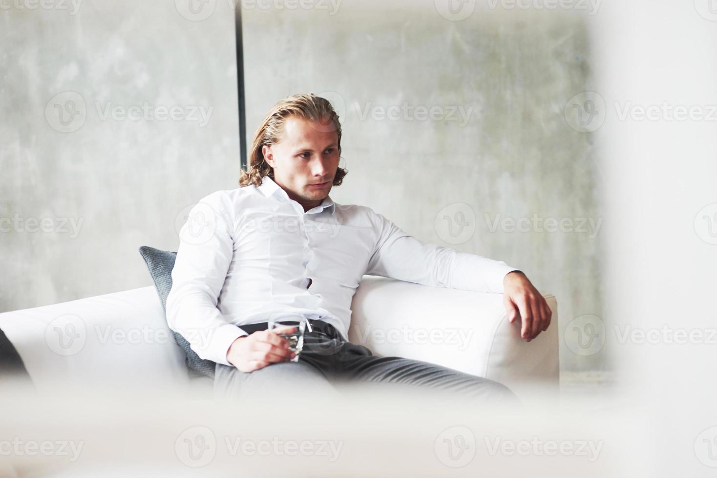 Serious young businessman sitting indoors on the sofa with whiskey glass photo