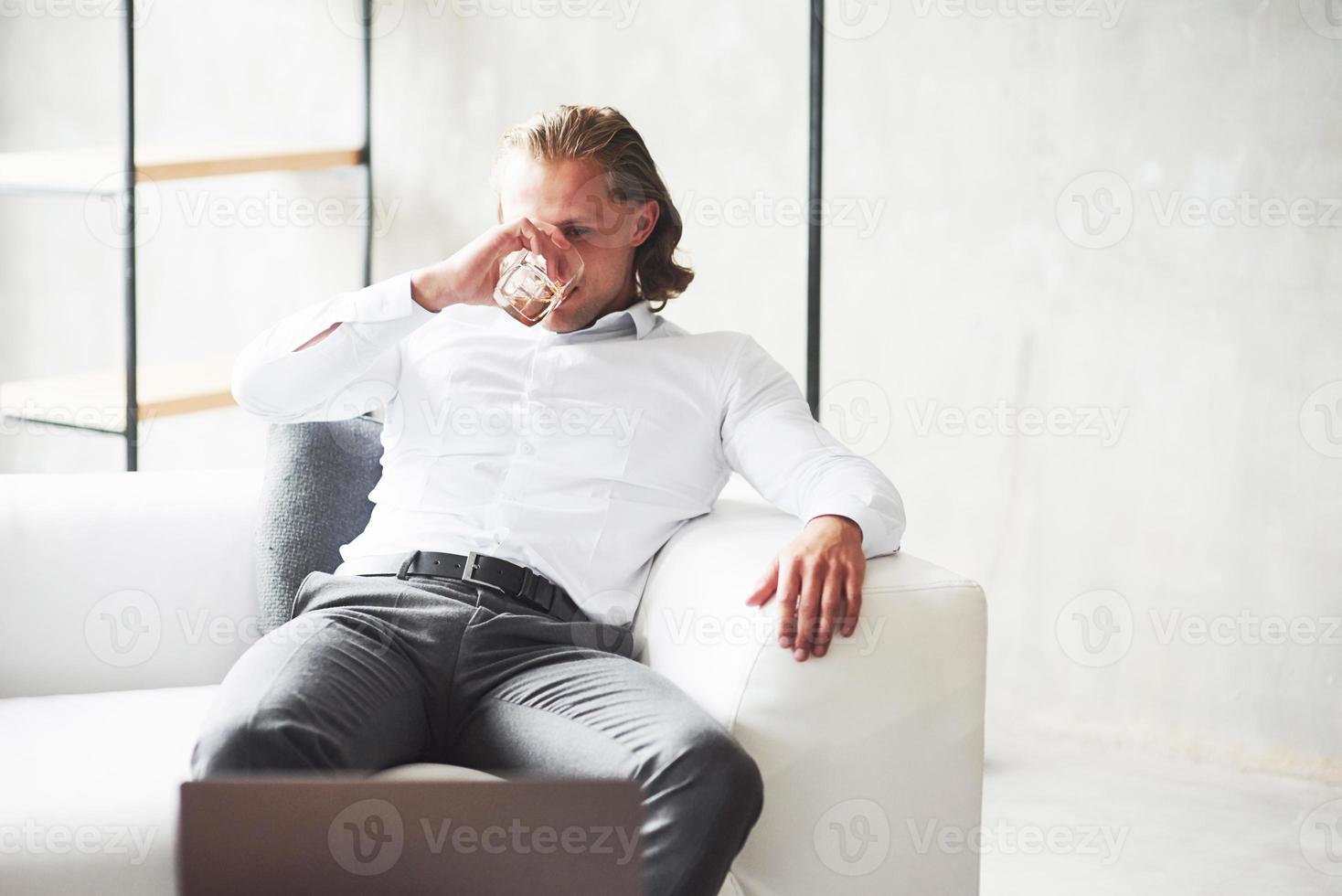 Young attractive office worker in classic wear sitting on the white sofa, drinking whiskey and looking at laptop photo