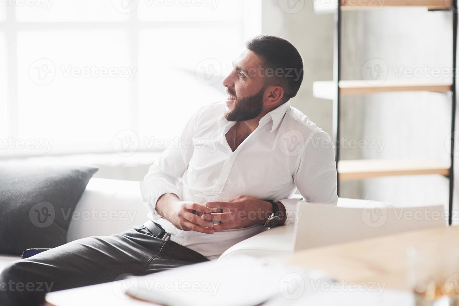 sonriendo mientras descansa. Foto de joven empresario barbudo con whisky en la mano sentarse en el sofá blanco