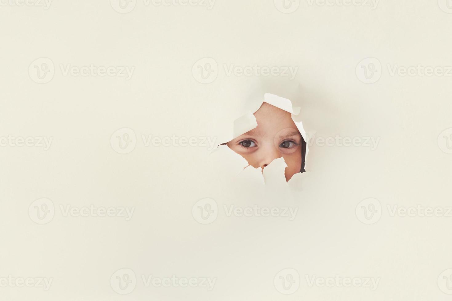 Spying game. Torn hole in white paper with eyes of little girl looking through photo