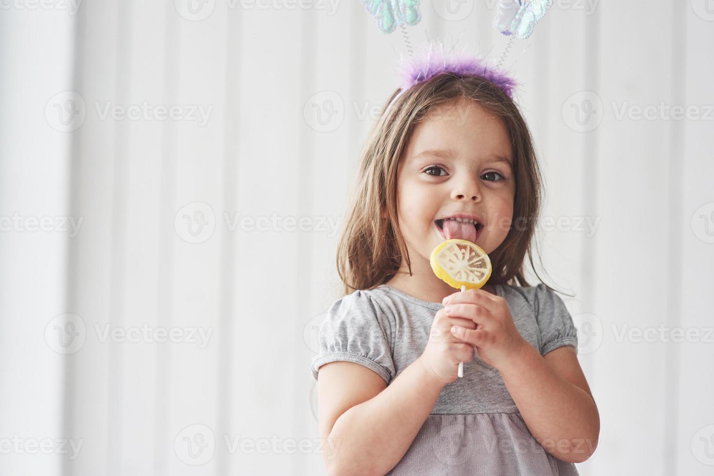 Niña bonita comiendo la vela amarilla con mariposas de juguete en la cabeza en la habitación blanca foto