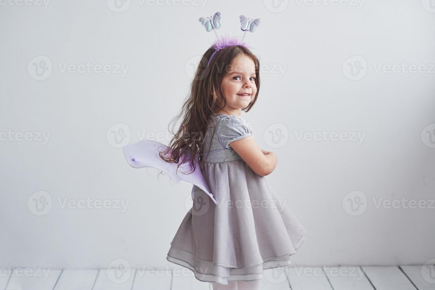 humor alegre y sonrisa sincera. Encantadora niña en traje de hadas de pie en la habitación con fondo blanco. foto