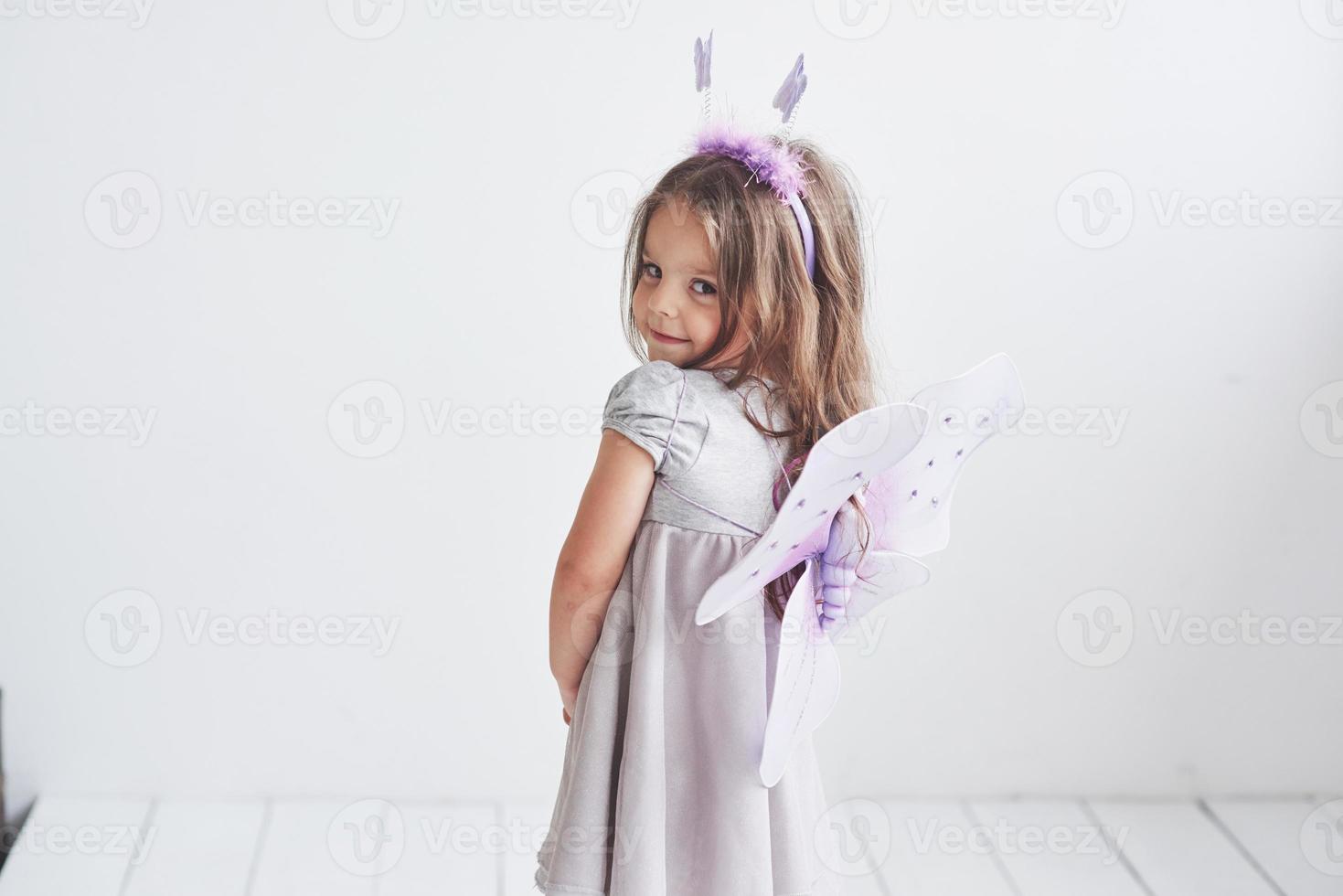 mirada tímida. Encantadora niña en el traje de hadas de pie en la habitación con fondo blanco. foto