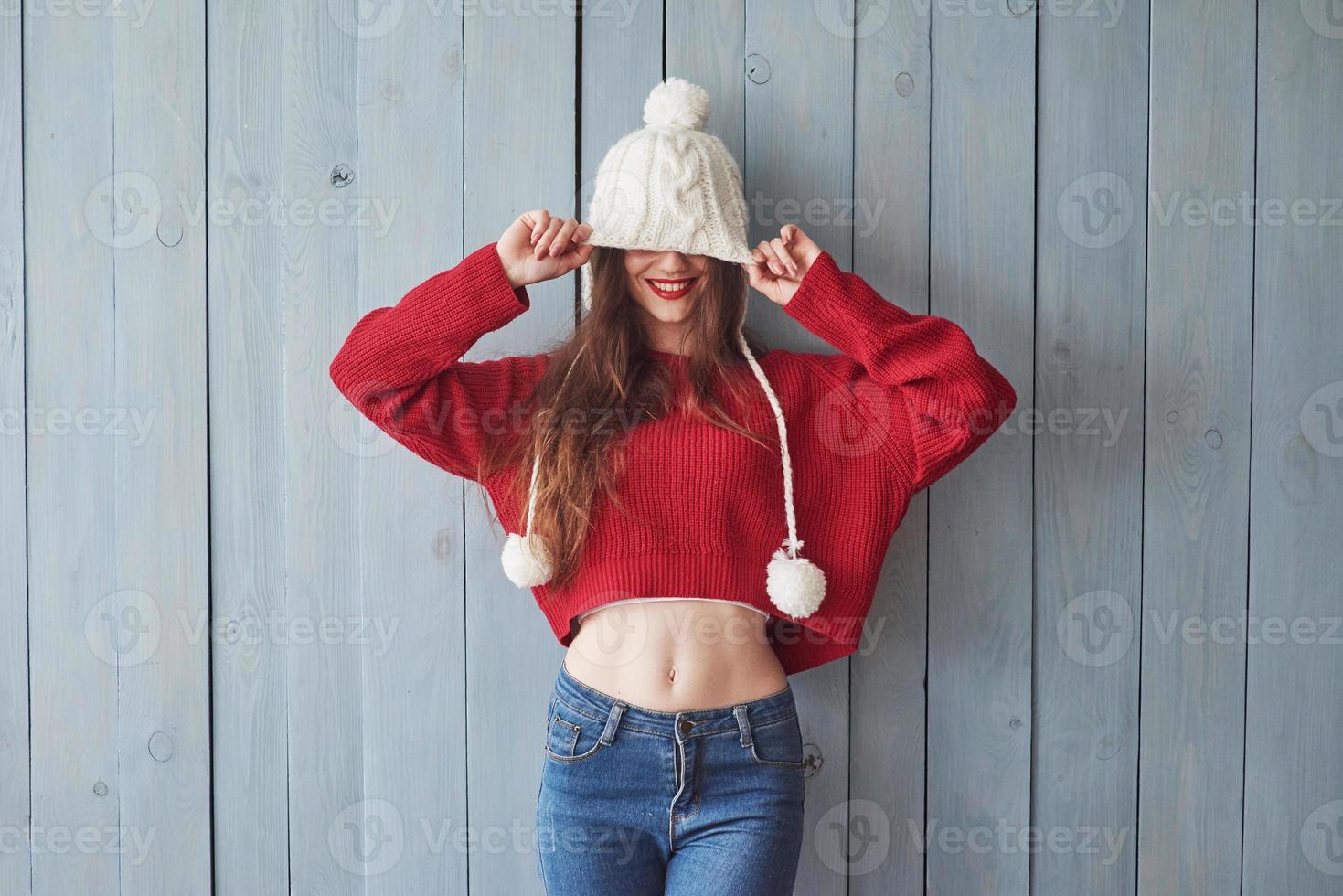 How long this thing is. It's covering my eyes. Funny girl in knitted sweater and hat standing indoors with wooden background photo