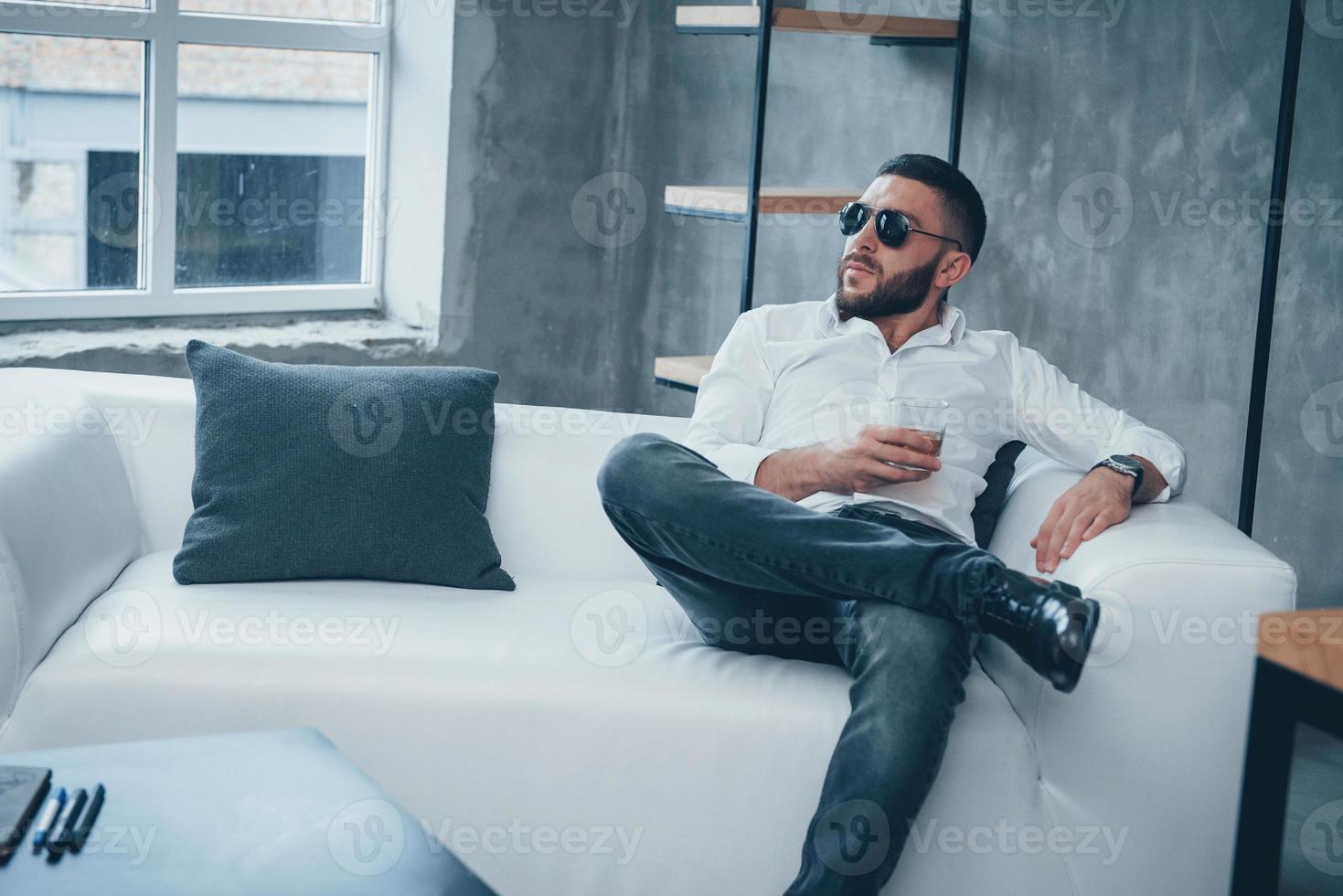 mirada seria pensativa. Joven hombre de pelo corto con gafas de sol sentado en un sofá en la oficina foto