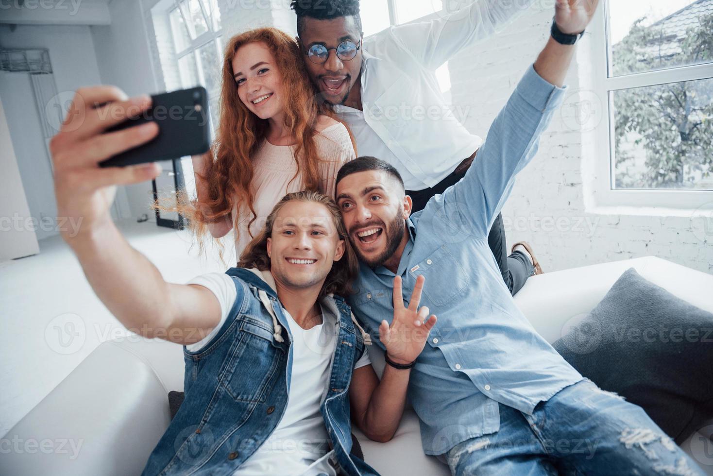 cara haciendo muecas. Alegres jóvenes amigos tomando autorretratos en el sofá y el interior blanco foto