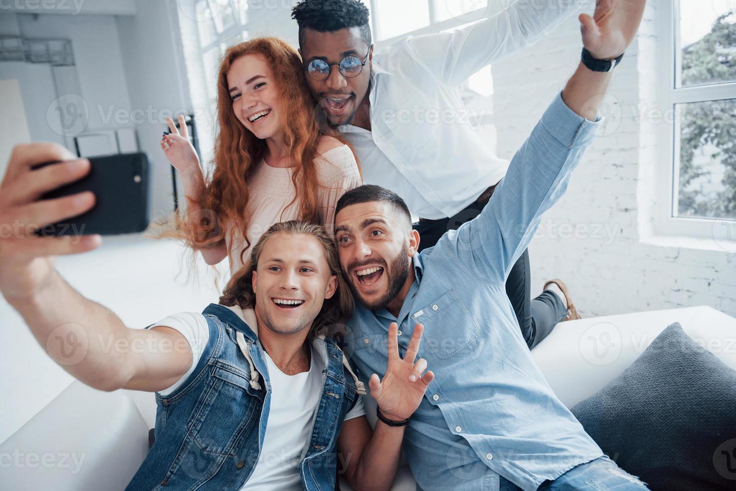 Such a lovely picture. Cheerful young friends taking selfies on sofa and white interior photo