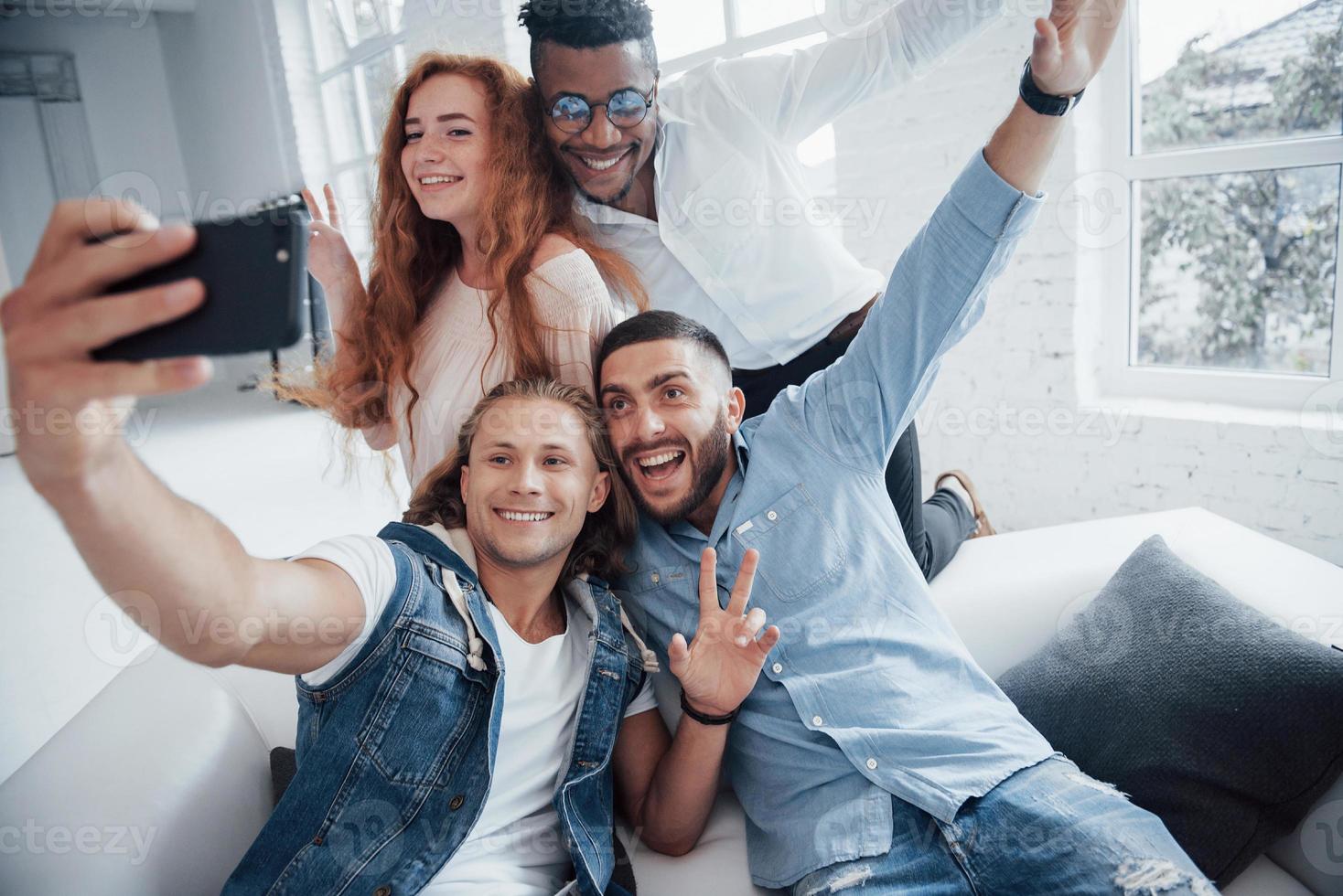 Muéstrame tus manos. Alegres jóvenes amigos tomando autorretratos en el sofá y el interior blanco foto