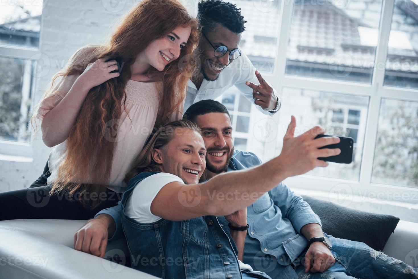 todo el mundo sonríe. Alegres jóvenes amigos tomando autorretratos en el sofá y el interior blanco foto