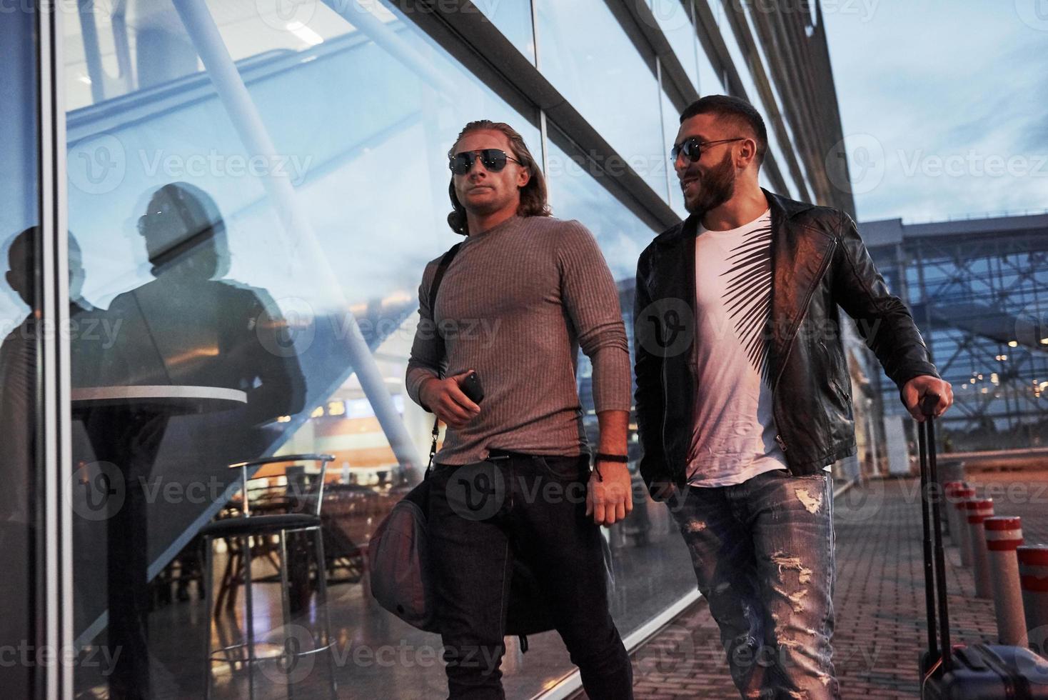 tener conversación mientras lleva equipaje. dos elegantes amigos con gafas de sol se han conocido cerca del edificio del centro de negocios y caminando frente a la cámara foto