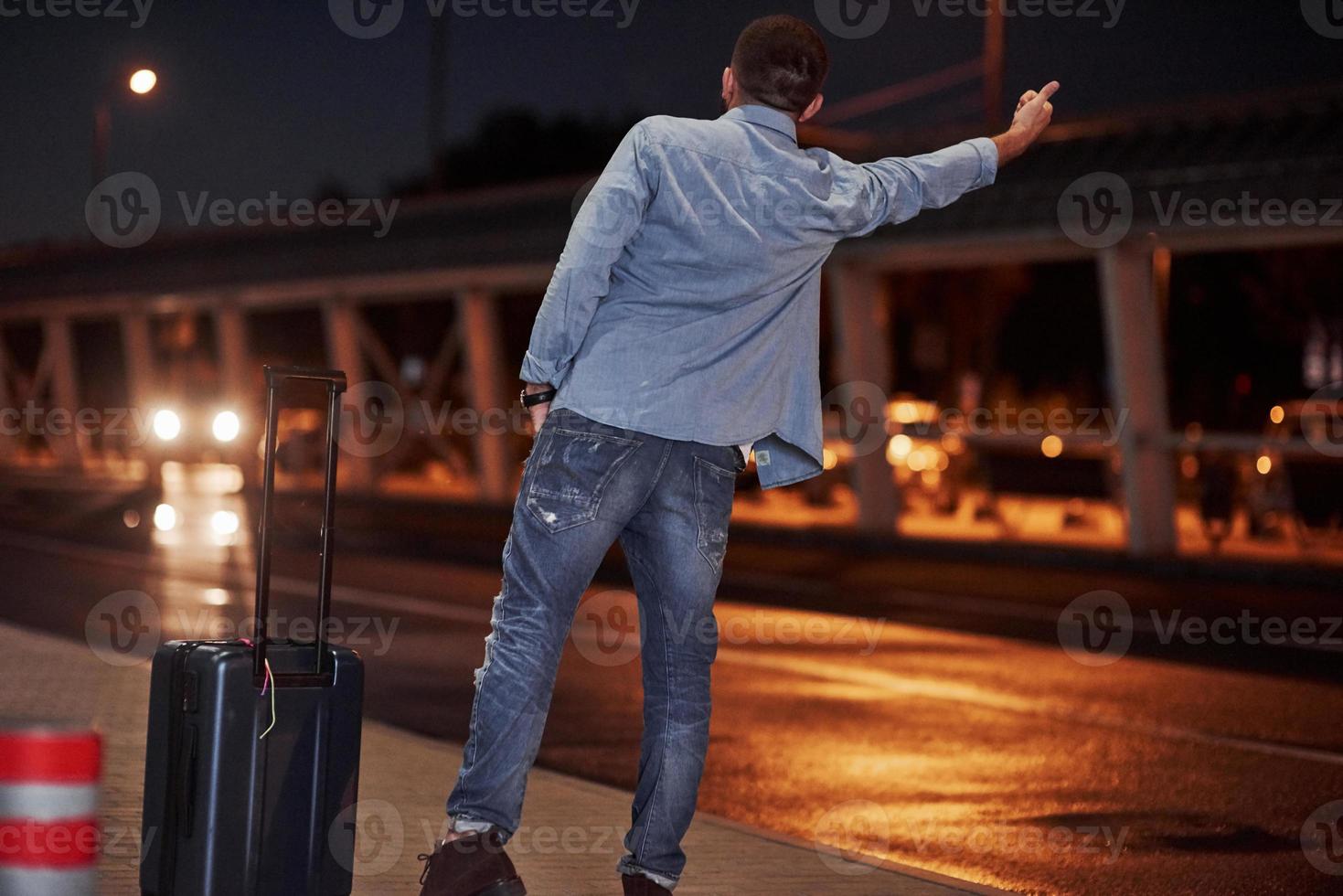 Unfortunately there is car at other side of the road. Short-haired man in jeans and a shirt with a baggage tries to catch the car photo