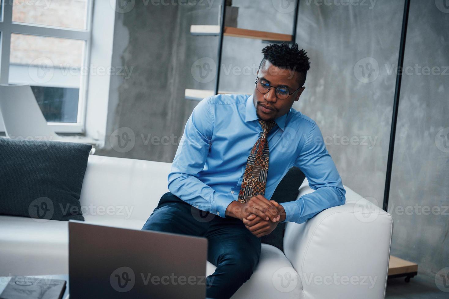 Foto de joven afroamericano con gafas y ropa clásica sentado en la parte delantera de la computadora portátil