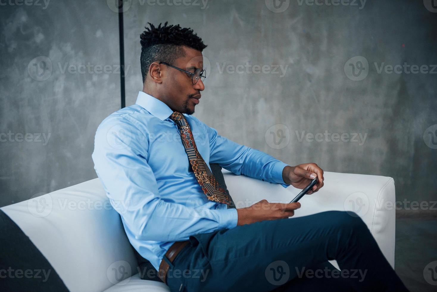 Concentraiting on the digital conversation. Close up portrait of young afro american guy in glasses and classic wear sitting on sofa photo