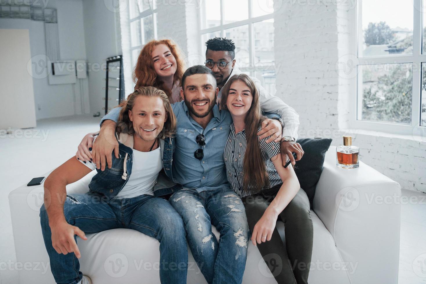 Divertidos jóvenes amigos divirtiéndose sentados en el sofá en el interior blanco foto
