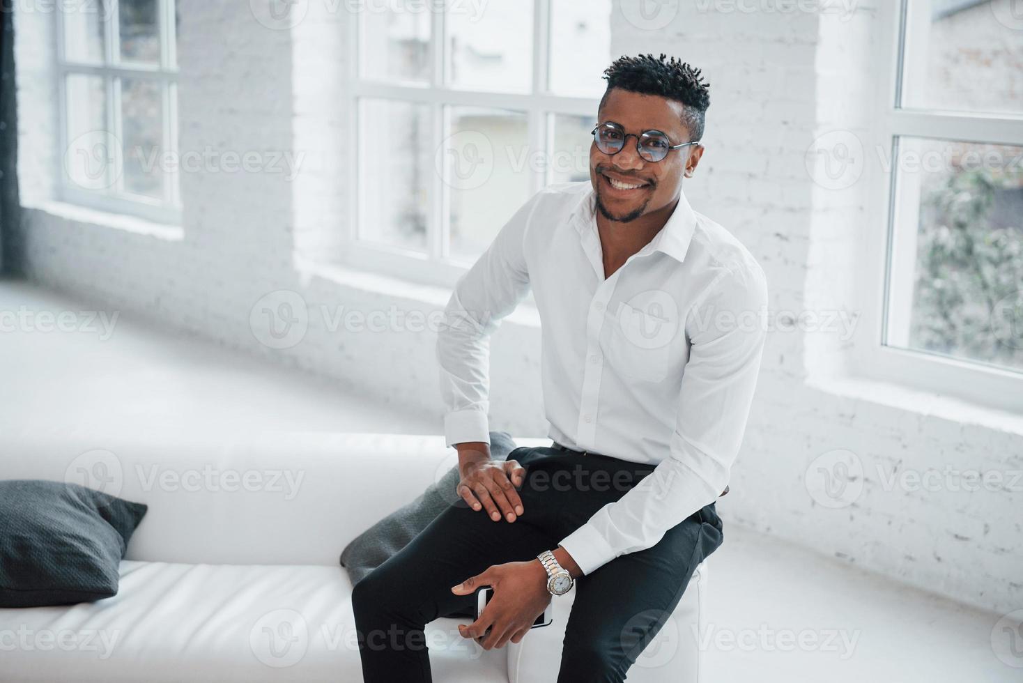 Elegante chico afroamericano en ropa de oficina y gafas sentado en el borde del sofá y sonriendo foto