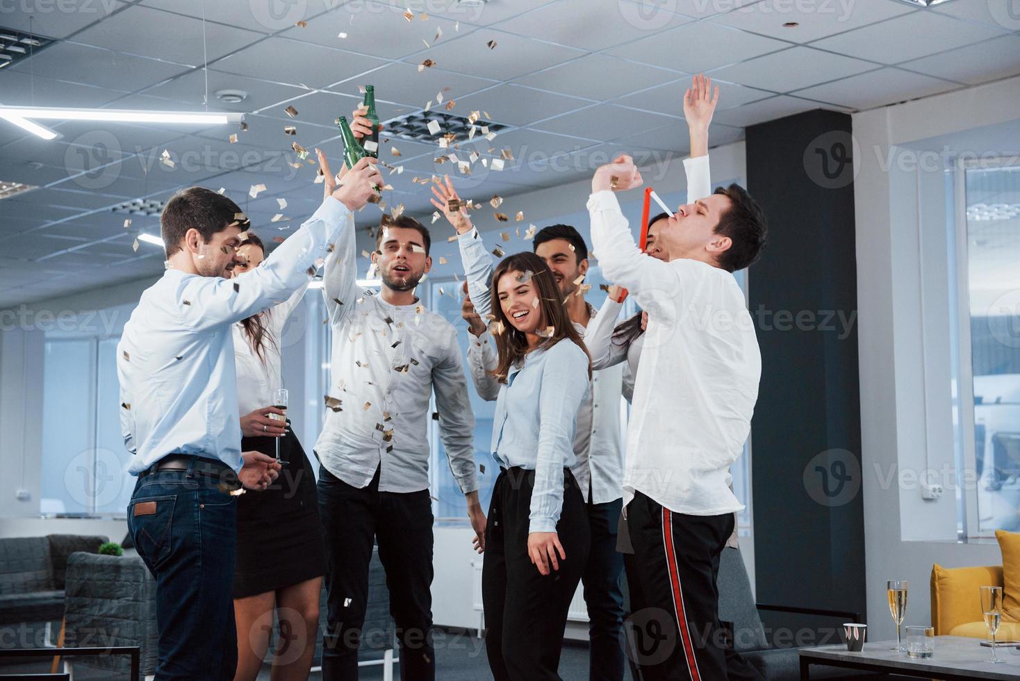 todos están felices. Foto de equipo joven en ropa clásica celebrando el éxito mientras sostiene bebidas en la moderna oficina bien iluminada