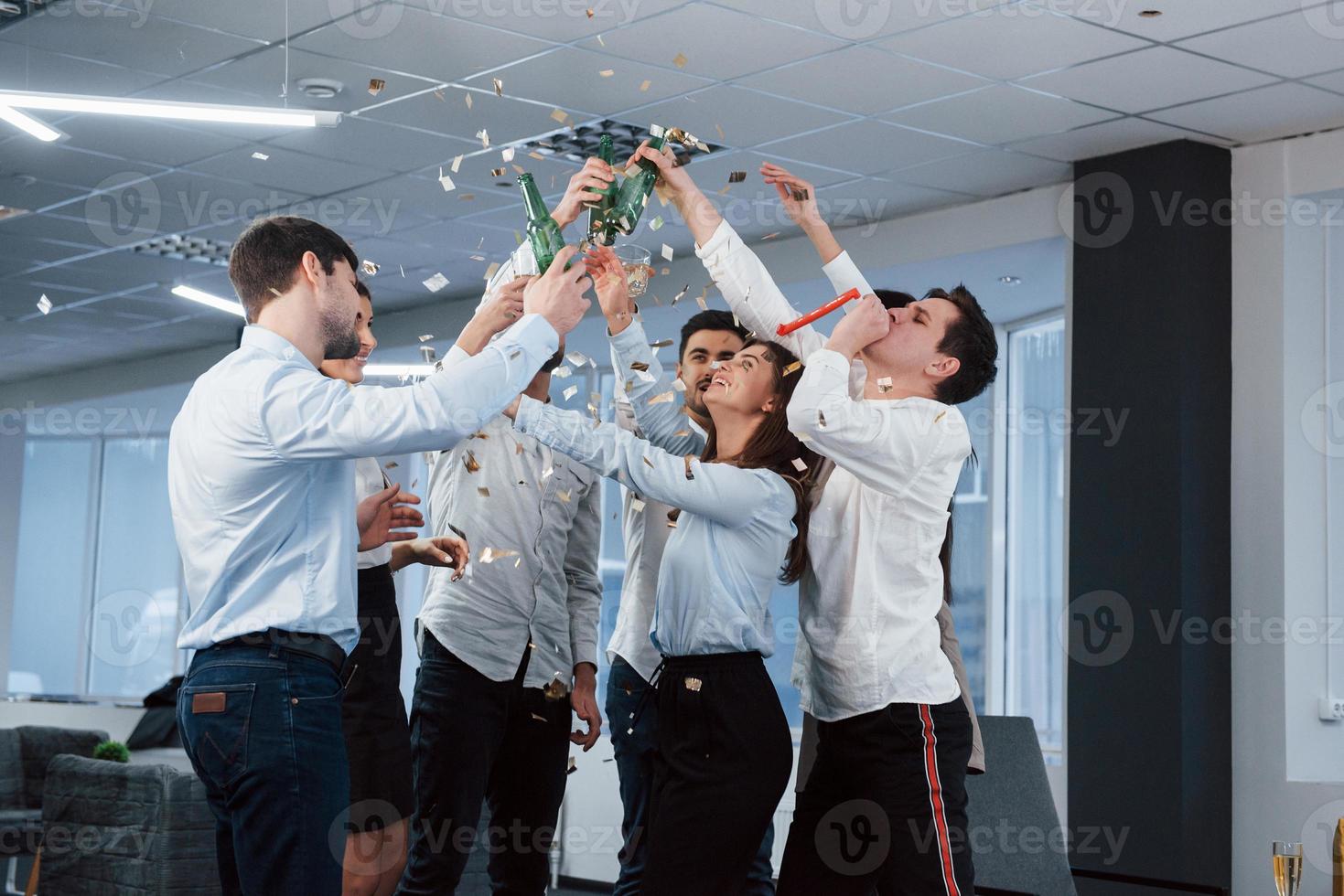 manos arriba. Foto de equipo joven en ropa clásica celebrando el éxito mientras sostiene bebidas en la moderna oficina bien iluminada