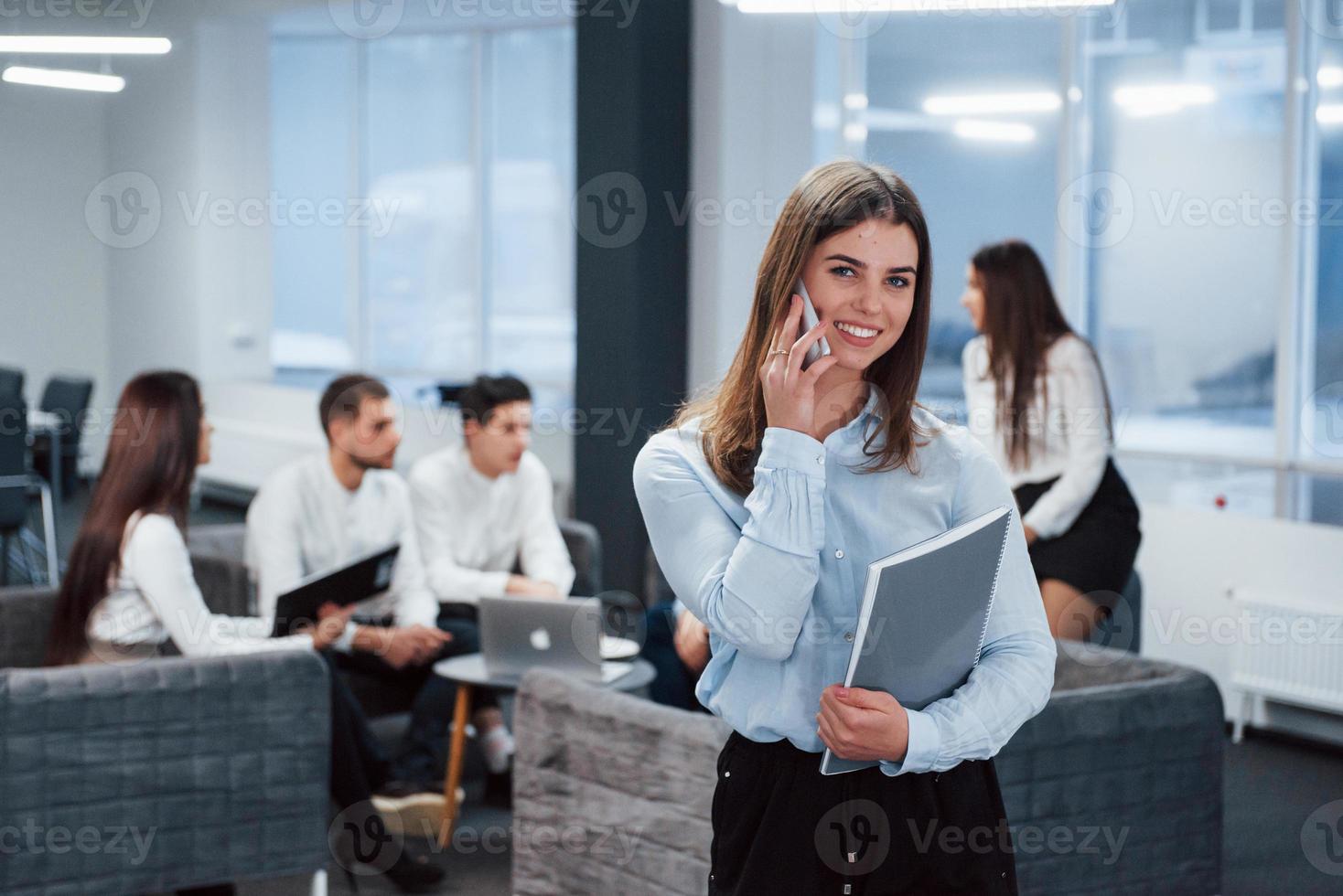 hablando por teléfono. Retrato de joven se encuentra en la oficina con empleados en segundo plano. foto