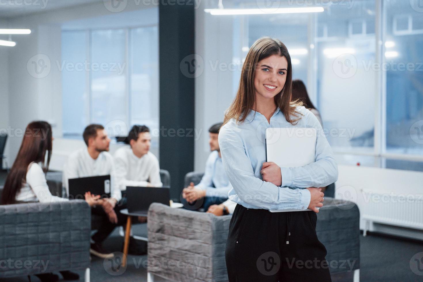 Classic wear. Portrait of young girl stands in the office with employees at background photo