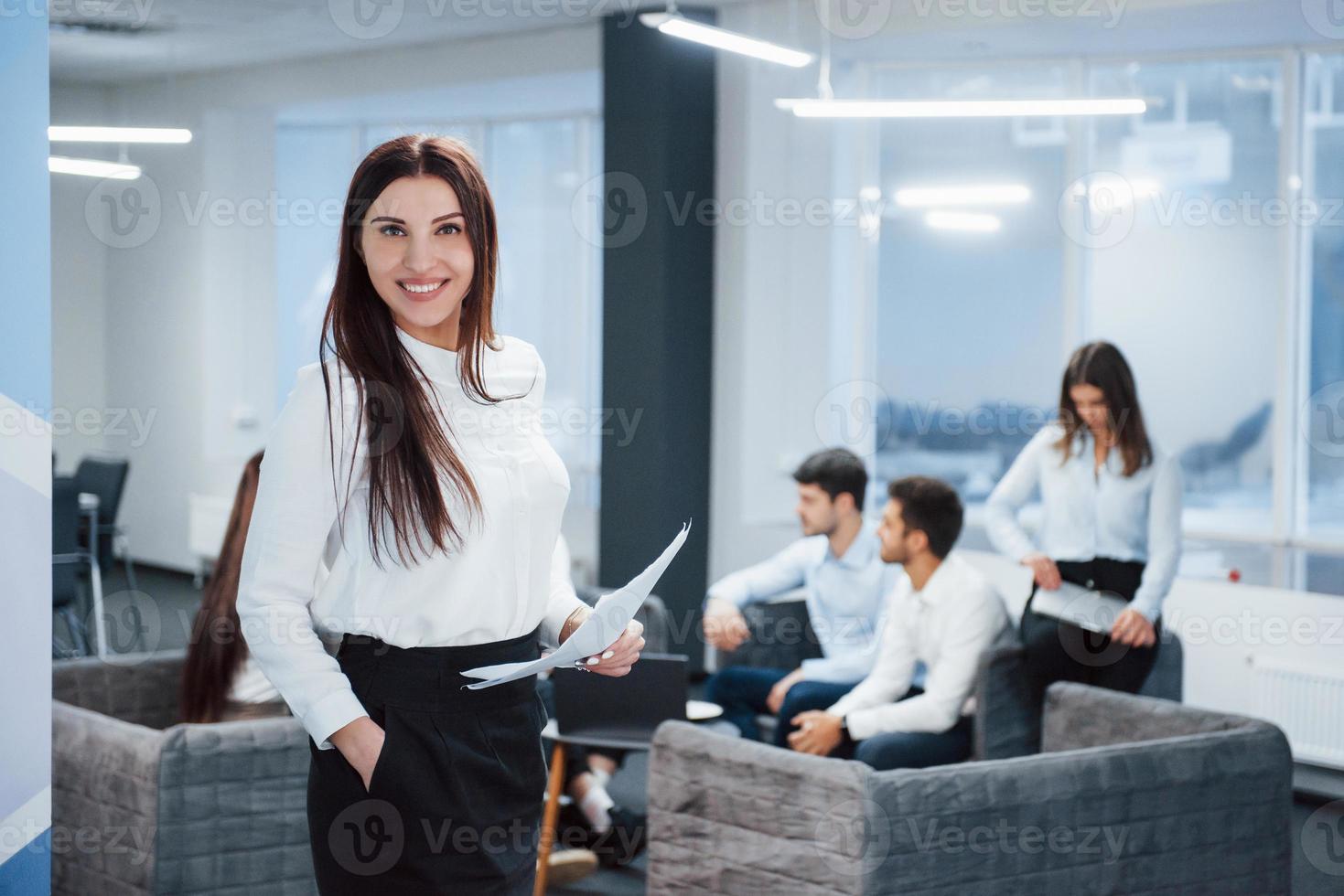 Friendly manager smiling to the camera. Portrait of young girl stands in the office with employees at background photo