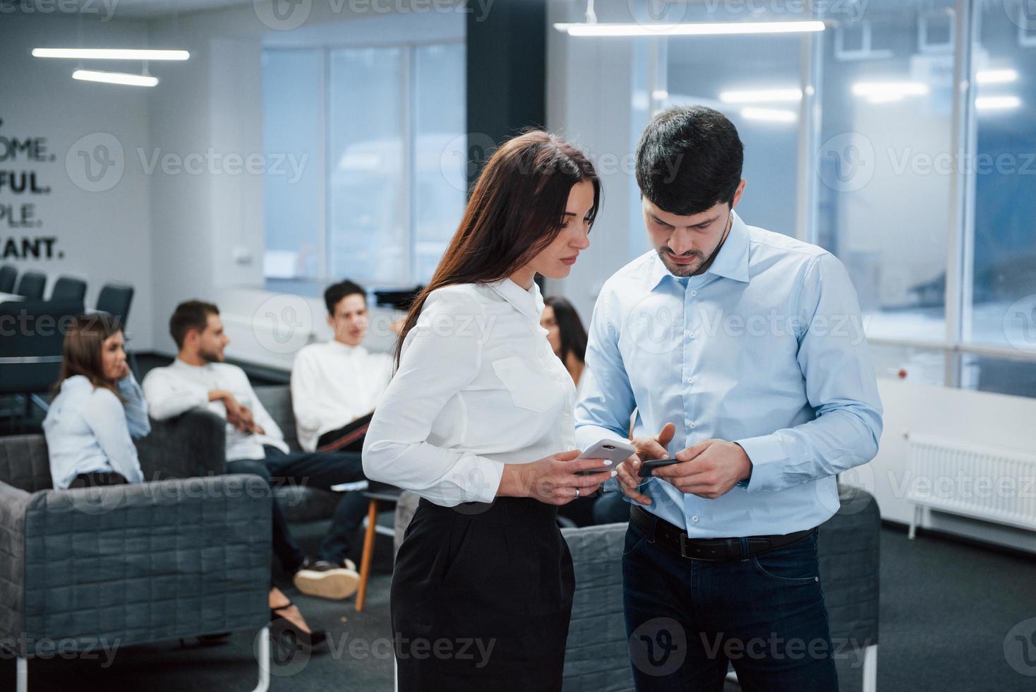 utilizando teléfonos inteligentes. Dos trabajadores de oficina con ropa clásica hablando frente a los empleados. foto