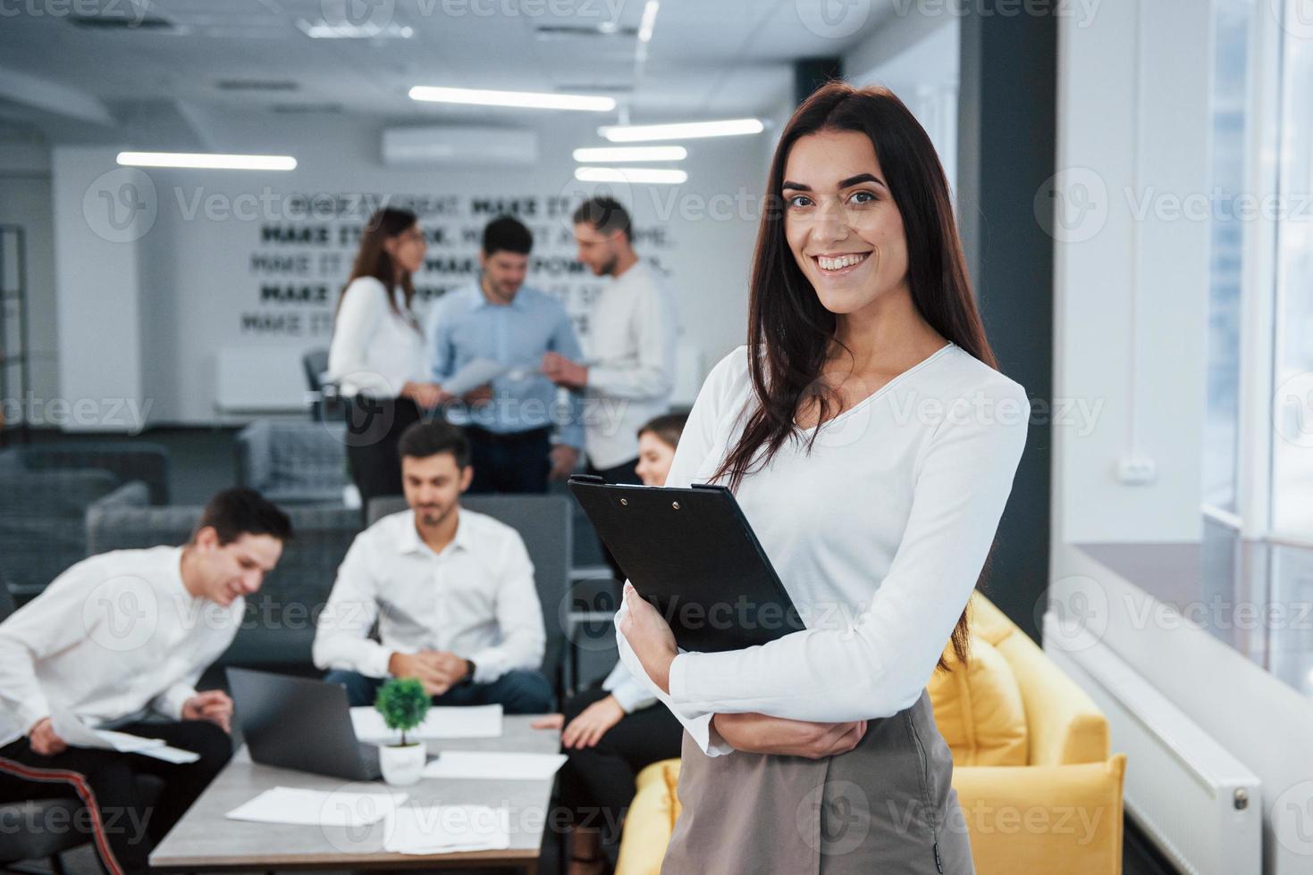 With notepad in hands. Portrait of young girl stands in the office with employees at background photo