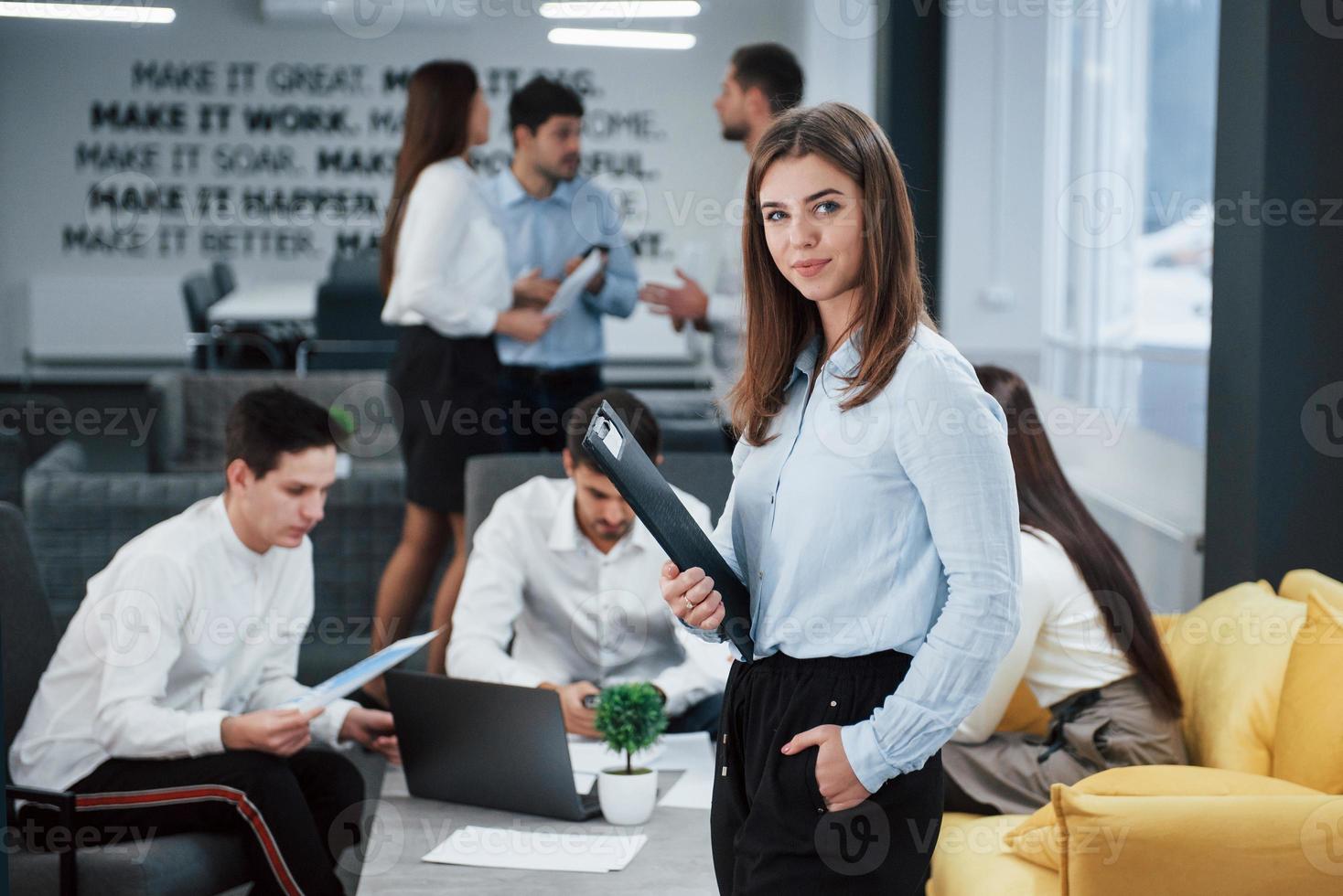 Turned sideways. Portrait of young girl stands in the office with employees at background photo