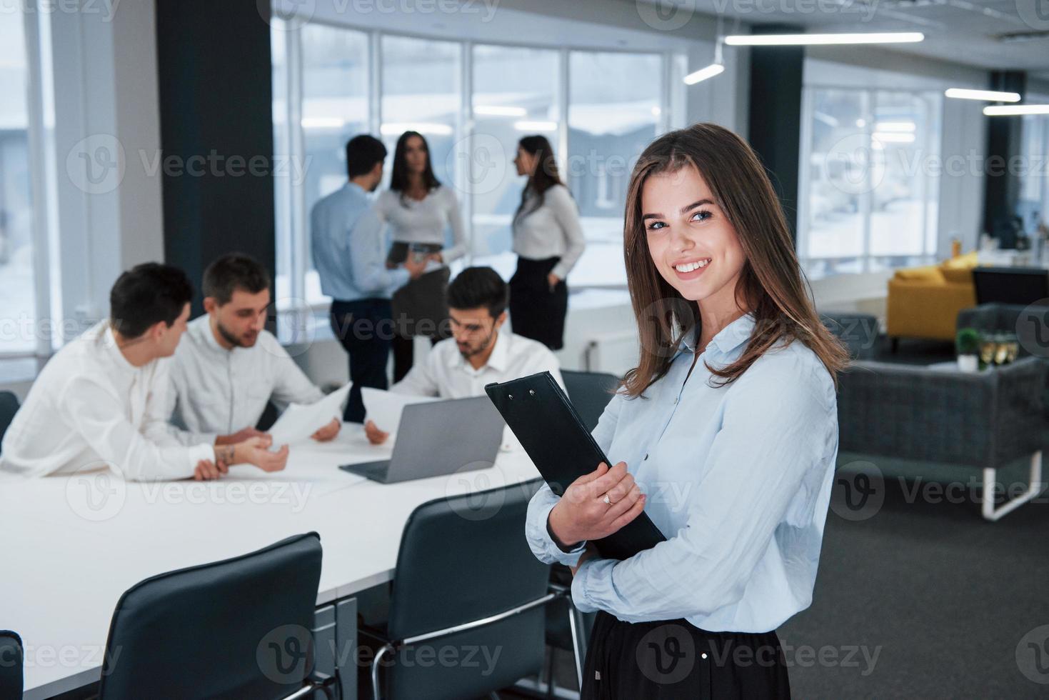 In white shirt and black pants. Portrait of young girl stands in the office with employees at background photo