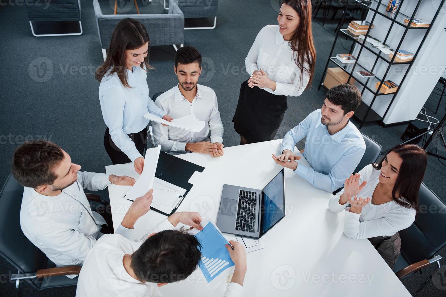 todo el mundo está de buen humor. Vista superior de los trabajadores de oficina en ropa clásica sentados cerca de la mesa usando una computadora portátil y documentos foto