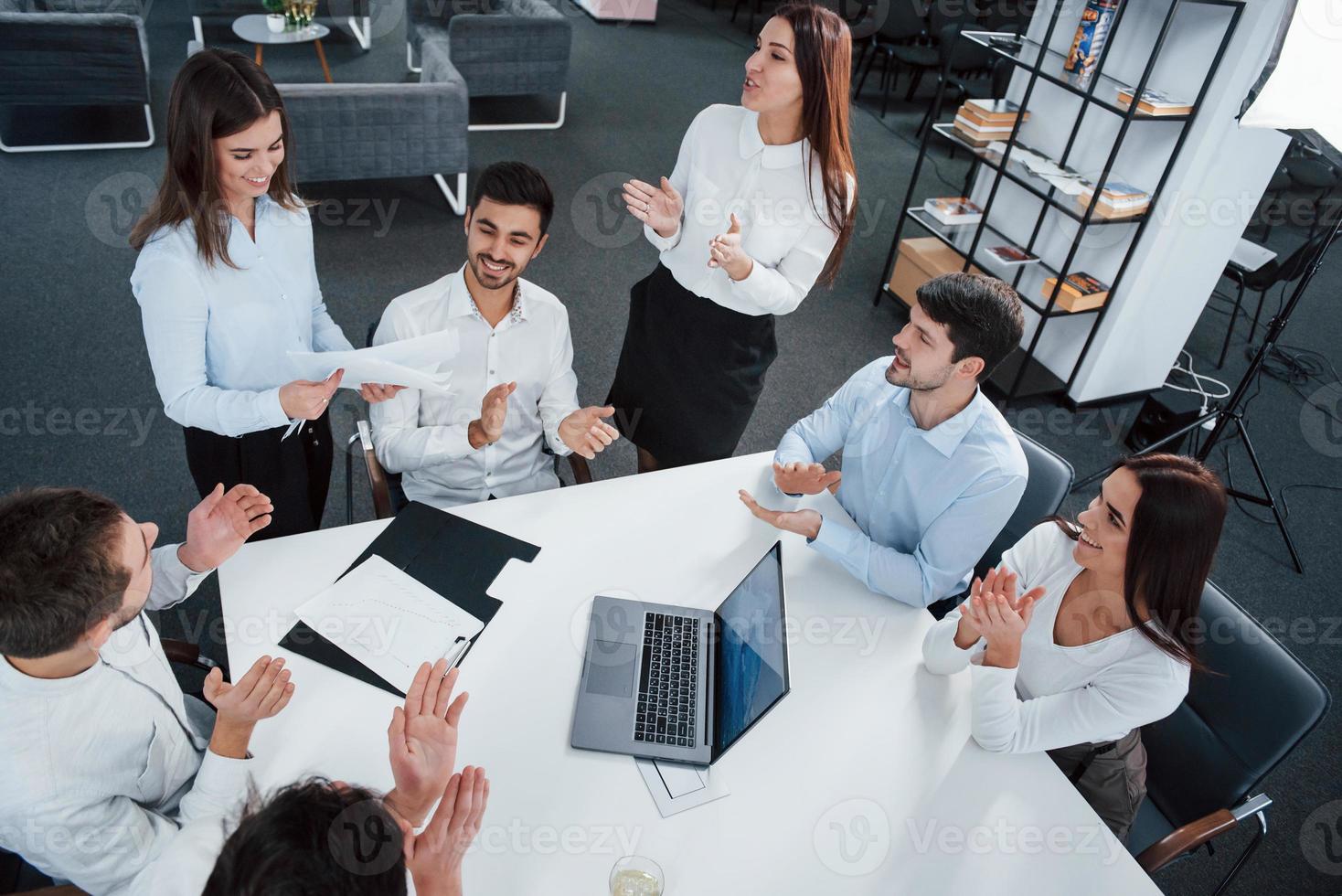 los empleados aplauden. Vista superior de los trabajadores de oficina en ropa clásica sentados cerca de la mesa usando una computadora portátil y documentos foto