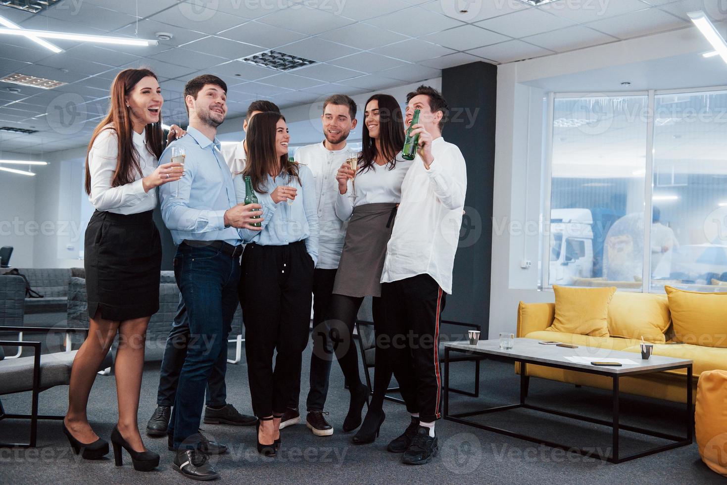 solo bromeando. Foto de equipo joven en ropa clásica sosteniendo bebidas en la moderna oficina bien iluminada