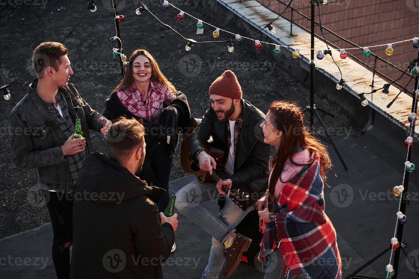 Vista superior de unos jóvenes amigos que beben, tocan la guitarra y se divierten en el techo con bombillas decorativas de colores foto