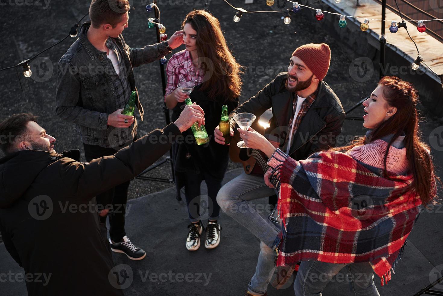 Party in full swing. Top view of a young friends that drinking, playing guitar and have fun at the roof with decorative colorful light bulbs photo