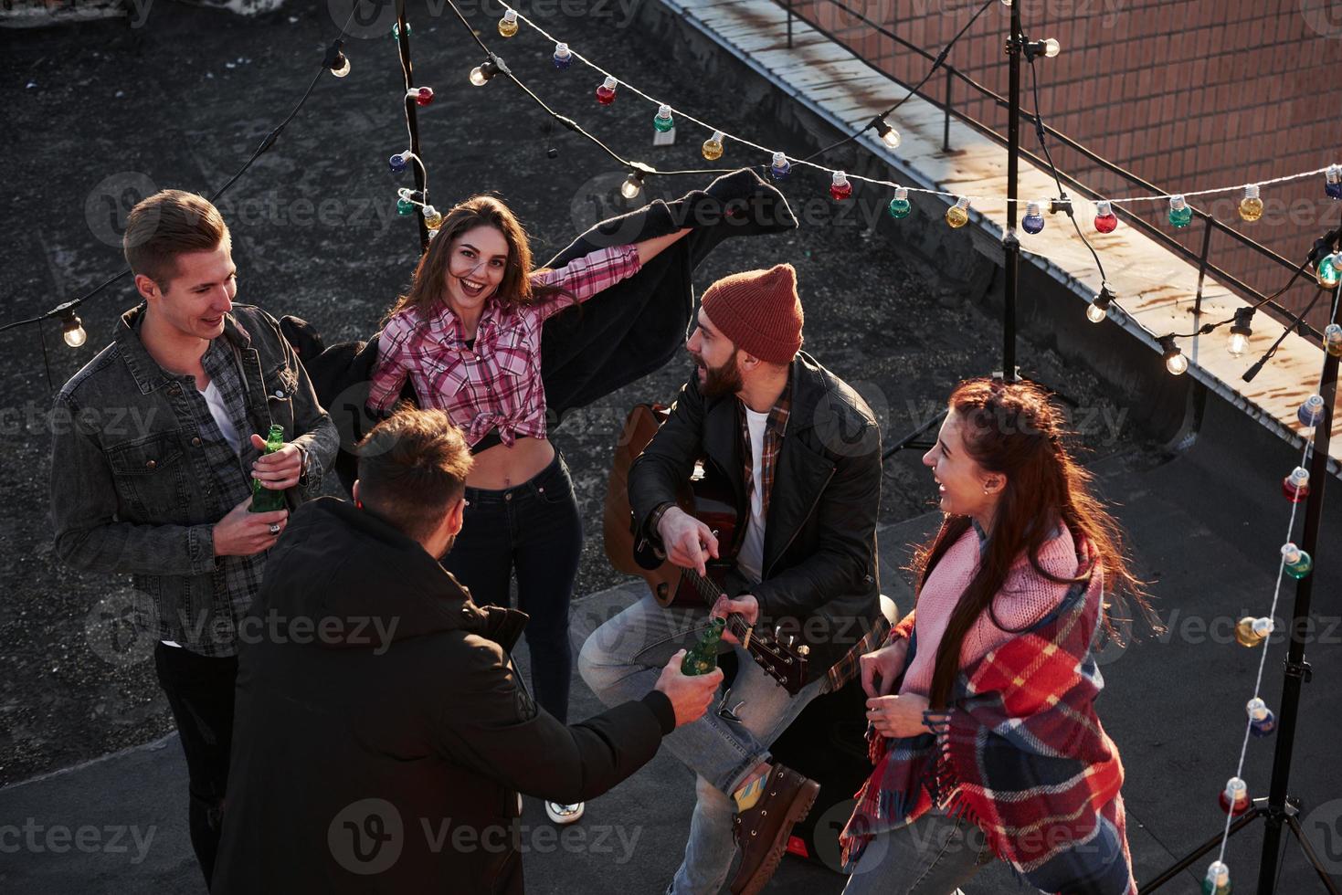 Attractive brunette girl smiles and posing for the picture. Top view of a young friends that drinking, playing guitar and have fun at the roof with decorative colorful light bulbs photo