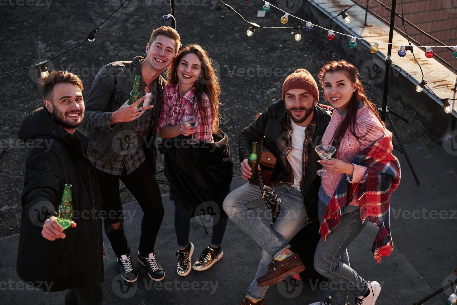 se abrazan y sonríen. gente positiva. Vista superior de unos jóvenes amigos que beben, tocan la guitarra y se divierten en el techo con bombillas decorativas de colores foto