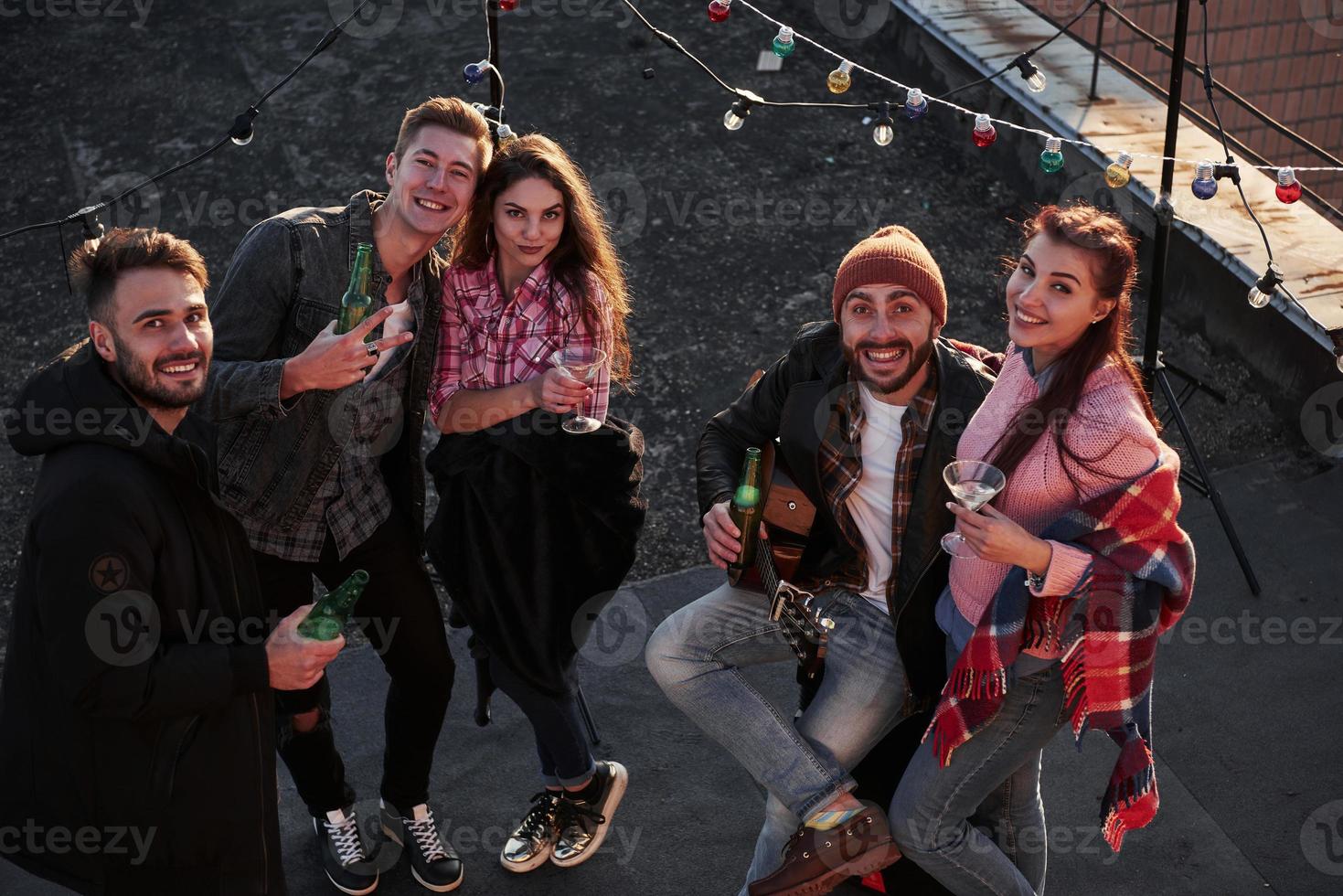 Looking up in the camera. Top view of a young friends that drinking, playing guitar and have fun at the roof with decorative colorful light bulbs photo